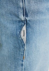 Helle blaue Denim-Jeans mit einer subtilen Textur, versehen mit einem aufgenähten Markenetikett an der Seitennaht und orangefarbenen Nähakzenten.
