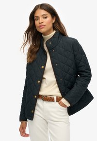 Femme portant une veste noire matelassée sur un pull en col roulé crème, un pantalon blanc et une ceinture tressée marron, se tenant avec une main dans la poche.