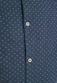 Dark blue shirt with a small, circular white pattern. Features white buttons and a textured fabric. Close-up on button placket.