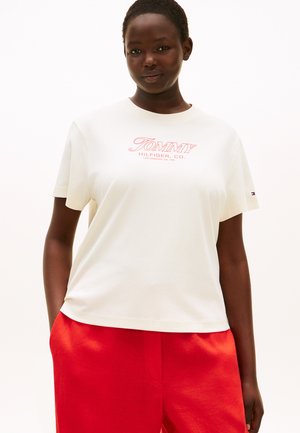 Ženska, ki nosi belo majico Tommy Hilfiger z rdečim logotipom in svetlo rdeče hlače, stoji z rokami v žepih pred belim ozadjem.