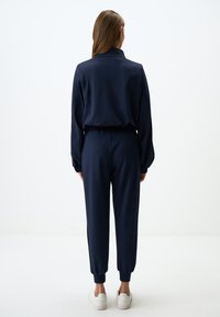 Tuta blu navy con una maglia colletto e polsini elasticizzati, caratterizzata da una vestibilità rilassata e pantaloni affusolati, abbinata a sneakers bianche.