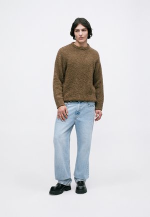 Pull en tricot texturé brun, coupe décontractée, col rond. Un jean bleu clair à jambes larges et des chaussures noires à semelles épaisses complètent un look décontracté.