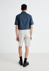 Camisa de manga corta con patrón en teal oscuro, pantalones cortos ligeros beige, zapatos negros y calcetines negros. Vista desde la parte de atrás, contra un fondo simple.