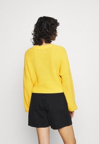 Suéter de punto amarillo con un corte cuadrado y costuras de hombro caídas, combinado con pantalones cortos de mezclilla negra. Tejido texturizado, diseño sencillo.