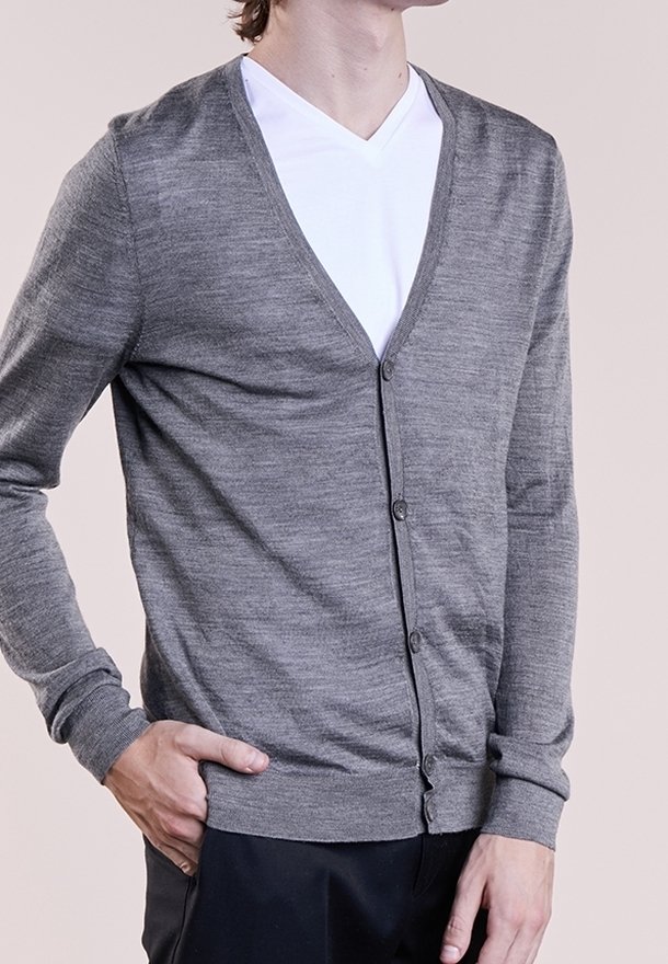 Mann trägt eine graue, geknöpfte Strickjacke über einem weißen V-Ausschnitt-Shirt, mit einer Hand in der Tasche und neutralem Hintergrund.