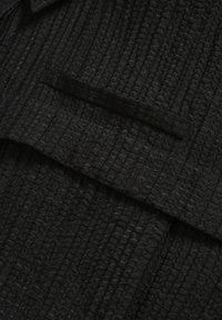 Indumento nero testurizzato con superficie arricciata. Presenta una tasca e un design a strati, enfatizzando un'estetica strutturata e minimalista.