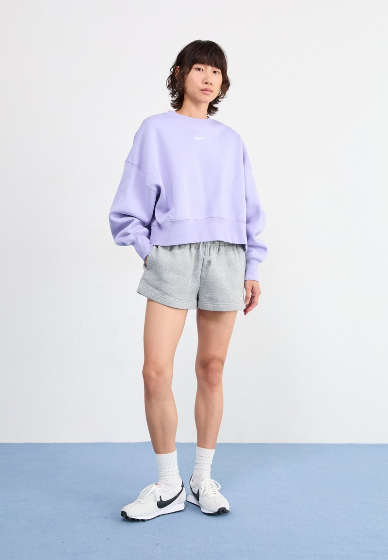 Sudadera recortada de color púrpura claro con pantalones cortos grises a juego. Logo de Nike en blanco en el pecho. Modelo lleva zapatillas blancas y calcetines blancos texturizados.