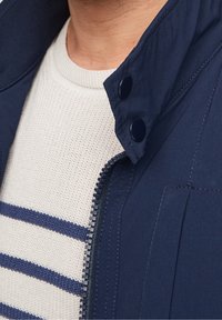 Veste bleu marine avec un col montant, une fermeture éclair et deux boutons-pression. Sous la veste, un pull crème à rayures bleues et tricot texturé.