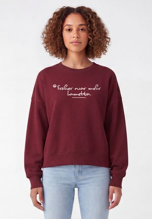 Femme aux cheveux bouclés portant un sweat bordeaux avec un texte allemand blanc et un jean bleu clair, debout devant un fond blanc uni.