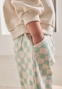 Hellbeiger Pullover mit Sprenkeln, kombiniert mit grün-cremefarben karierten Hosen. Die Hosen verfügen über Seitentaschen und eine weiche Stoffstruktur.