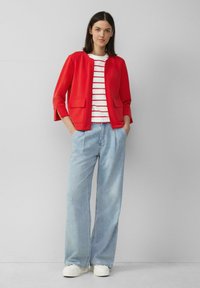 Rode, gestructureerde blazer met een ronde halslijn en twee voorzakken, gedragen over een witte gestreepte shirt. Gecombineerd met lichtblauwe wijde jeans en witte sneakers.