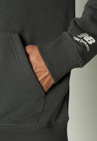 Felpa grigia scuro con polsini a coste e tasca frontale. Presenta un logo "New Balance" bianco sulla manica, con una texture morbida.