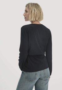 Blonde Frau mit kurzem Haar, die ein schwarzes Langarmoberteil und eine blaue Jeans trägt, steht mit dem Rücken zur Kamera und schaut zur Seite.