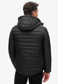 Giacca a vento nera con cappuccio, caratterizzata da strisce orizzontali trapuntate e una texture liscia. Cappuccio regolabile con dettagli a cordino.
