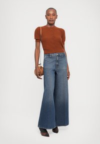 Brown short-sleeve cardigan with buttons, paired with wide-leg blue denim jeans. Model holds a small crossbody bag. Black pointed heels.