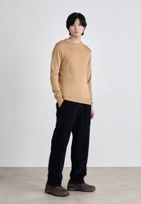 Beiger Langarmpullover, Rundhalsausschnitt, weicher Stoff; kombiniert mit dunklen, locker sitzenden Pants und braunen Sportschuhen mit grauen Sohlenakzenten.