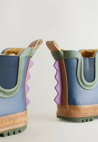 Bunte Gummistiefel mit blauen, lila und grünen Abschnitten, strukturierten Sohlen und einem dekorativen braunen Band mit orangefarbener Schrift.