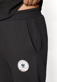 Black sweatpants with a smooth texture, featuring side pockets and a circular logo with the German Football Association in white and colors beneath.