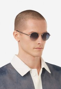 Ümmargused päikeseprillid tumeda gradientläätsete, õhukese hõbedase raami ja minimalistliku disainiga. Mudel kannab kontrastset kraega särki.