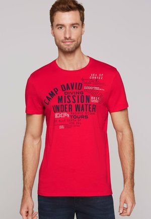 Mann trägt ein rotes T-Shirt mit schwarz-weißen tauchbezogenen Texten und steht vor einem schlichten hellgrauen Hintergrund.