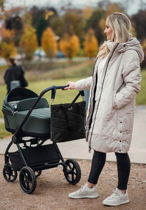 Kinderwagen mit einer dunkelgrünen Babywanne und schwarzem Rahmen, begleitet von einer schwarzen, gesteppte Wickeltasche. Eine Person in einem hellen, gesteppte Mantel hält den Kinderwagen.