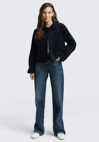 Jonge vrouw met lang haar in een zwarte knoopjack, blauwe high-waisted jeans en witte sneakers, staande tegen een effen achtergrond.