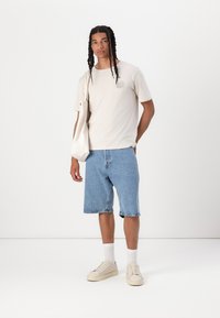 Tricou din bumbac bej cu un grafic circular pe piept, asortat cu pantaloni scurți din denim de culoare albastră deschisă. Tenisi albi și șosete până la gleznă finalizează ținuta.