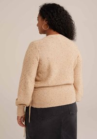 Bež pulover z zlatimi pikami, strukturni pletenina, dolgi rokavi in prilegnjen pas. Ima vrvice na hrbtni strani za prilagodljivost.