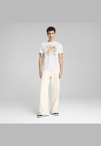 Camiseta blanca con gráfico de zapatillas amarillas, combinada con pantalones cremosos holgados y zapatillas en blanco y negro. Fondo neutro.