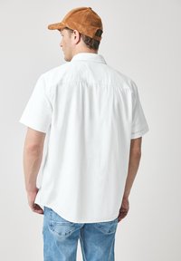 Witte korteblazende shirt met een rechte snit en een zachte textuur, gecombineerd met blauwe denim jeans. Een bruine suède baseballpet wordt gedragen.