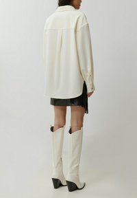 Witte, oversized button-up shirt met een achternaad, gecombineerd met een zwarte minirok met franjes en kniehoge witte laarzen met hak.