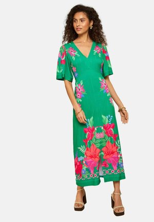 Žena s kučeravými vlasmi nosí zelené midi šaty s ružovým a červeným kvetinovým vzorom, výstrih do V, krátke rukávy, zlaté šperky a sandále na platforme.