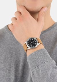 Silbernes Analog-Armbanduhr mit schwarzem Zifferblatt, großen weißen Zahlen und braunem Wildlederarmband. Getragen an einer Hand mit einem grauen Pullover.