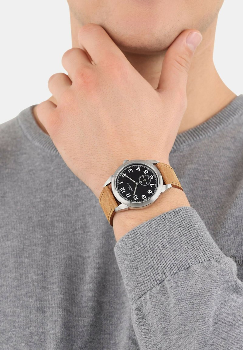 Silbernes Analog-Armbanduhr mit schwarzem Zifferblatt, großen weißen Zahlen und braunem Wildlederarmband. Getragen an einer Hand mit einem grauen Pullover.