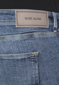 Un bolsillo trasero de unos vaqueros de mezclilla azul con una etiqueta de color marrón cosida encima, que muestra el nombre de la marca "RIVER ISLAND" en texto negro.