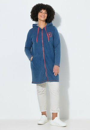 Kvinde smilende med krøllet hår iført en blå hætteklædt lang jakke med lynlås og røde detaljer, hvide bukser og hvide sneakers.