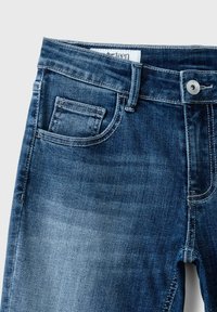 Gros plan sur un jean en denim bleu montrant les passants de ceinture, la poche avant, la petite poche, le bouton métallique et la texture décolorée du tissu.