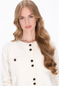 Crème-kleurige gebreide cardigan met een geribbelde halslijn, zwarte knopen en een borstzak aan de linkerkant. Zachte textuur, casual pasvorm en knoopaccenten.