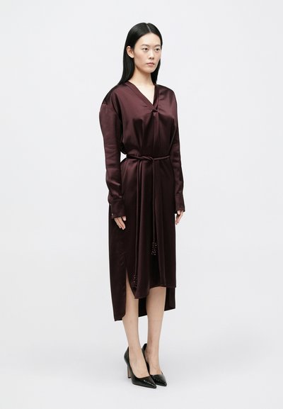 Φόρεμα με σατέν ύφασμα στο χρώμα της μπορντό, μακρυά μανίκια, βαθύ V-λαιμόκοψη, πλάγιες σχισμές και ζώνη στη μέση. Φορώντας μαύρα παπούτσια με τακούνι.