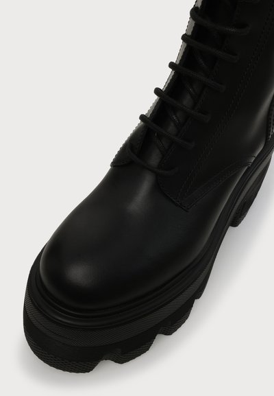 Botte de combat en cuir noir avec une semelle épaisse et texturée et des lacets noirs, vue en plongée inclinée sur un fond clair.
