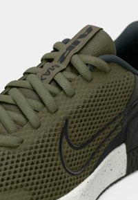 Olivově zelené sportovní boty s síťovinovým svrškem, plochými tkaničkami a černým logem Nike. Texturovaný design se skvrnitou bílou mezipodrážkou a tmavou podrážkou.