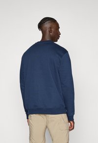 Man met een gevlochten kapsel, gekleed in een marineblauwe sweatshirt en beige cargo-broeken, staand met de rug naar de camera tegen een effen lichte achtergrond.
