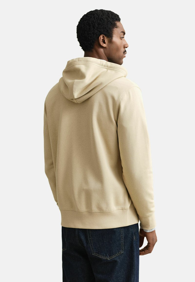 Mann trägt beige Kapuzenpullover und dunkelblaue Jeans, steht seitlich mit hauptsächlich sichtbarem Rücken vor einfarbigem Hintergrund.