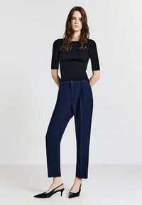 Schwarzes, geripptes Oberteil mit kurzen Ärmeln, kombiniert mit hochgeschnittenen marineblauen Hosen und schwarzen Slingback-Pumps mit spitzer Zehenform. Glatte Texturen und figurbetonende Silhouette.