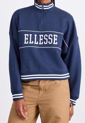 Personne portant un sweat-shirt bleu marine à fermeture quart de zip avec des rayures blanches et le texte "ELLESSE", associé à un pantalon beige.