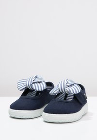 Zapatillas de lona azul marino con una suela de goma blanca, que cuentan con un detalle de lazo rayado en azul y blanco en la parte delantera y un diseño de punta abierta.