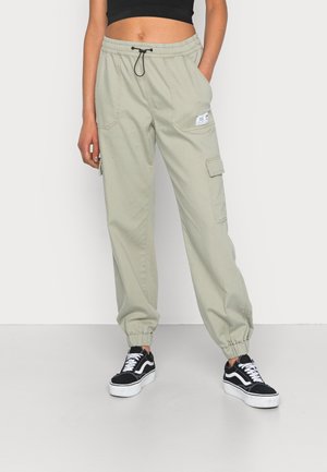 Jasnoszare spodnie cargo z elastycznymi mankietami i talią, czarnym sznurkiem, bocznymi kieszeniami, noszone z czarno-białymi trampkami i czarnym krótkim topem.