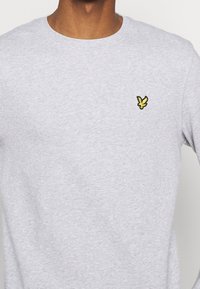 Sudadera gris con cuello redondo, que presenta un pequeño logo de mariposa bordado en negro y amarillo en el lado izquierdo del pecho. Textura suave.