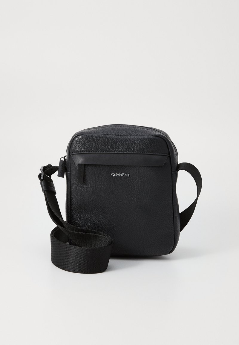 Borsa a tracolla in pelle nera di Calvin Klein, forma quadrata, superficie texturizzata, tracolla regolabile, tasca frontale con zip e branding minimale.