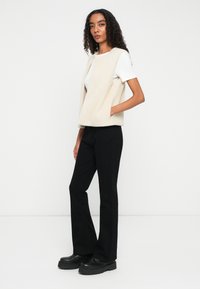 Eine Frau mit lockigem Haar steht vor einem weißen Hintergrund und trägt ein weißes kurzärmeliges Hemd, eine beige ärmellose Weste, schwarze Hosen und schwarze Plateauschuhe.